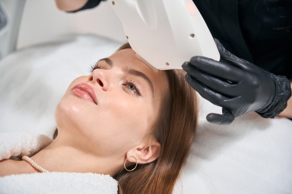 Cropped female cosmetologist doing laser depilation of face of woman on couch